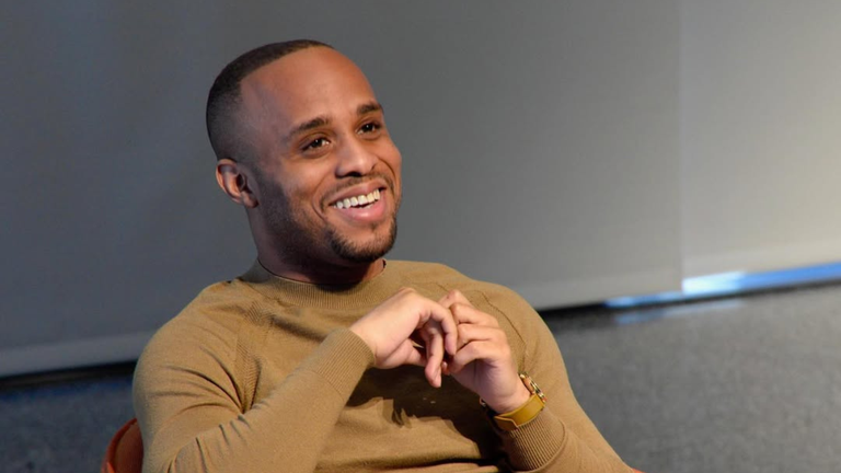 Christopher Gray, founder of Scholly, smiling while seated in a relaxed setting, wearing a tan sweater and a gold watch.