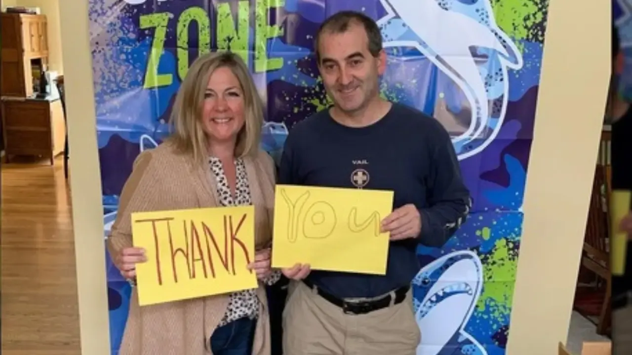 Patty and Andy Watne smiling and holding yellow thank-you signs, standing in front of a colorful shark-themed backdrop, expressing gratitude to customers after their Shark Tank episode.
