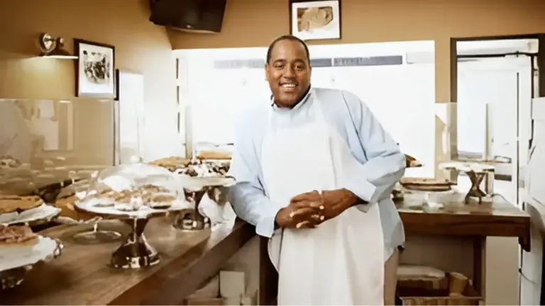 Tod Wilson, wearing a blue shirt and white apron, smiling while leaning on a counter filled with pies in his bakery.