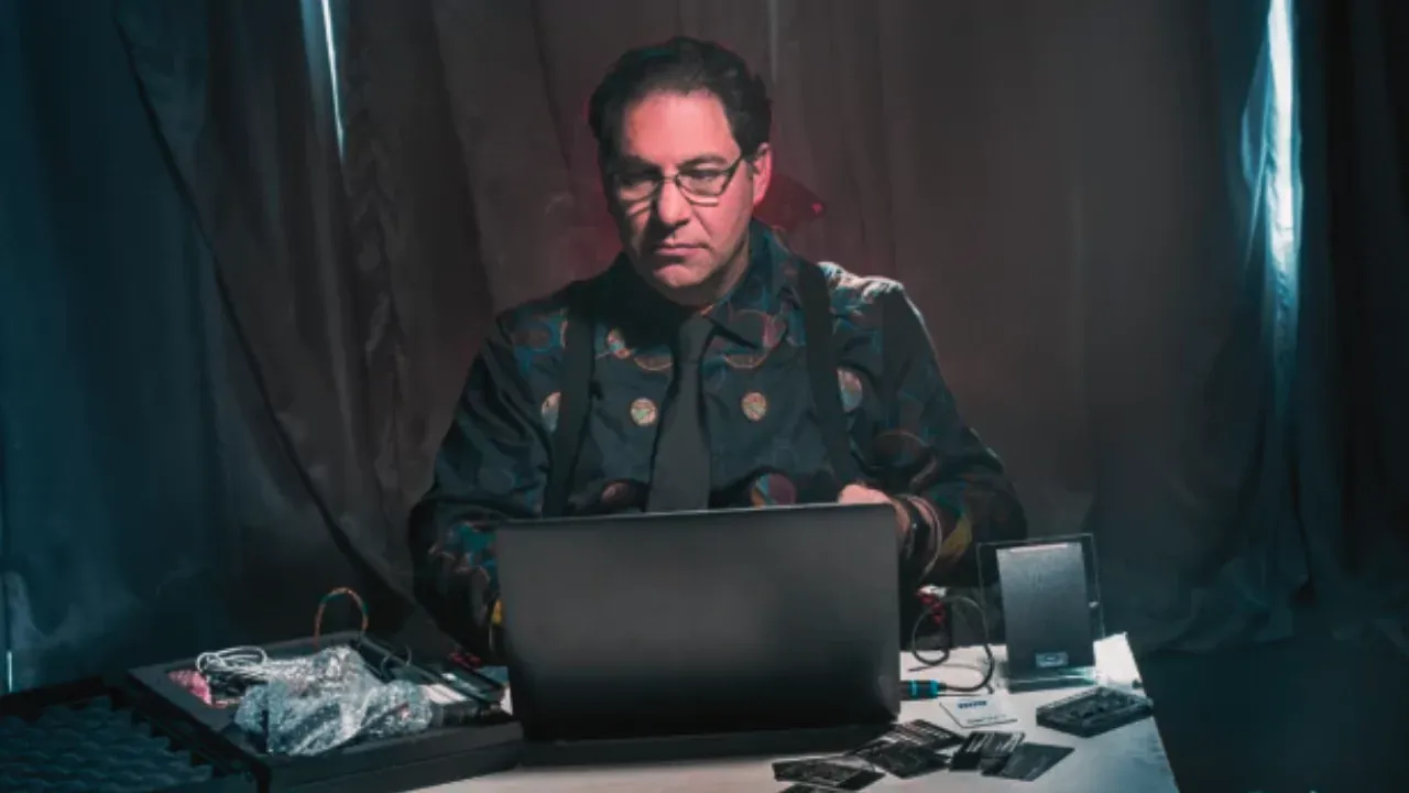 A serious man in a dark, patterned shirt sits at a desk with a laptop and hacking tools, representing Kevin Mitnick, once the world’s most wanted hacker.