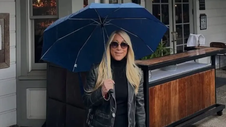 Lori Greiner holding a blue umbrella, wearing sunglasses and a leather jacket outside a restaurant.