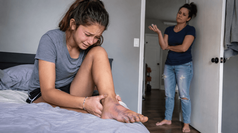 Teen girl crying over injured foot while her mother dismisses her pain.