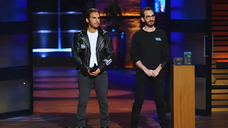 Two founders of Chubby Buttons stand on the Shark Tank stage, dressed in black, presenting their Bluetooth remote with a display stand beside them.